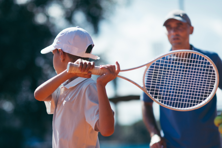 Ein Junge hält einen Tennisschläger, während ein Trainer im Hintergrund zusieht.