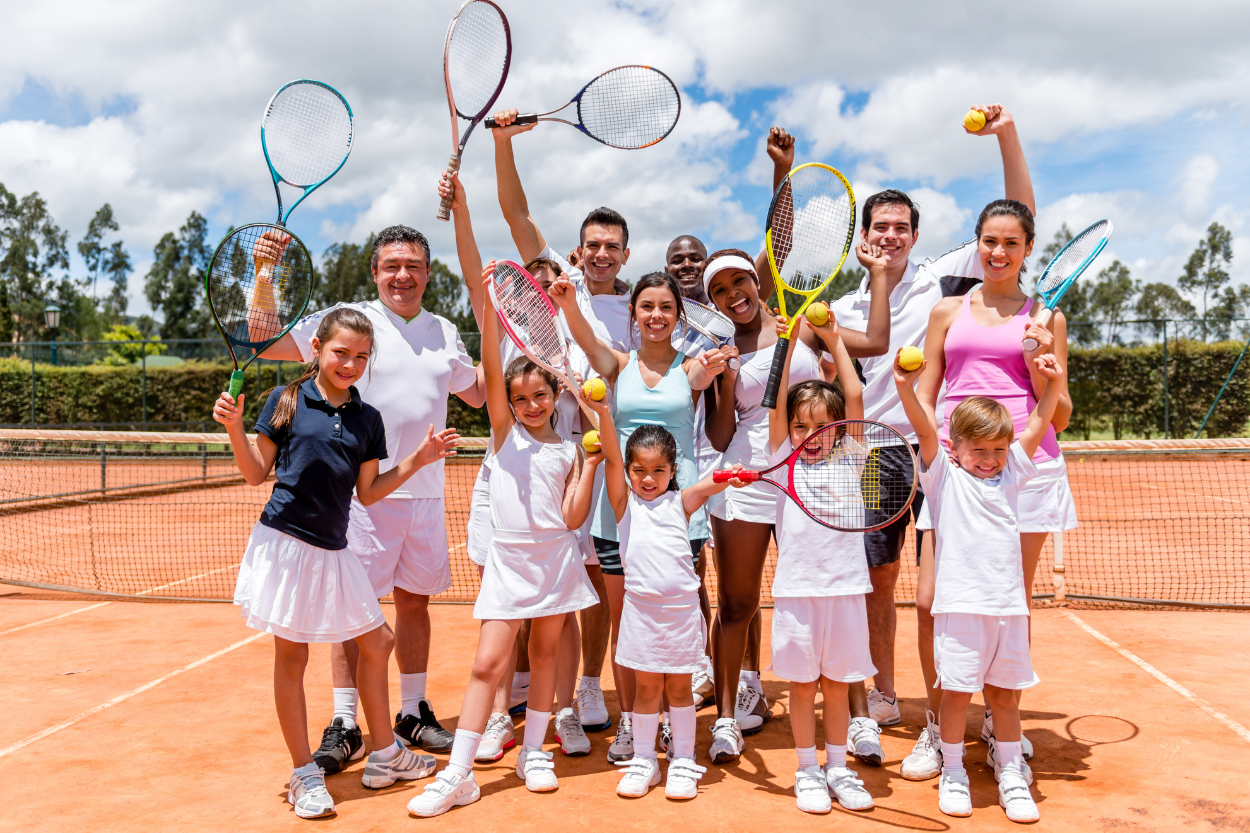 Gruppe von Erwachsenen und Kindern in weißen Outfits mit Tennisschlägern und Bällen auf dem Platz.